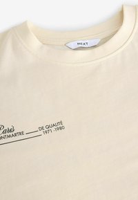 Tricou de culoare crem cu guler rotund. Prezintă text negru: "Paris Montmartre De Qualité 1971-1980". Textură din bumbac moale și neted.
