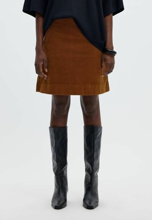 Personne portant une jupe marron jusqu'aux genoux, des bottes noires montant jusqu'aux genoux, un haut noir et un bracelet noir au poignet droit.