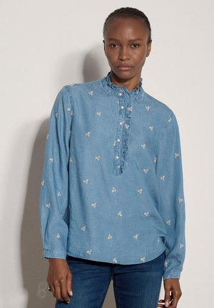 Mujer que lleva una blusa azul claro de manga larga con pequeñas flores blancas bordadas y cuello con volantes, combinada con vaqueros azul oscuro.