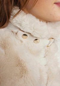 Col haut en fausse fourrure crème avec une texture douce, doté d'une sangle en tissu beige et de deux boutons-pression dorés pour la fermeture.