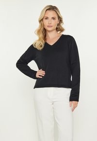 Pulover negru cu guler în V, fabricat dintr-un material tricotat moale, cu mâneci lungi și o croială lejeră, asortat cu pantaloni albi clasici.