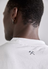 Camiseta blanca de algodón con un pequeño acento gráfico gris en la parte posterior, con cuello redondo y mangas cortas. Detalle del tejido texturizado visible.