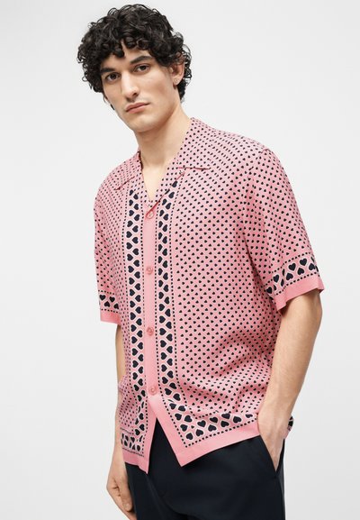 Νέος άνδρας με σγουρά μαλλιά, που φοράει ένα ροζ πουκάμισο με κοντά μανίκια και μαύρες καρδιές, και σκούρο παντελόνι, στέκεται μπροστά σε ένα απλό φόντο.