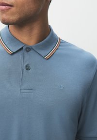 Ljusblå pikétröja med ett texturerat tyg, med en randig krage i grönt, brunt och vitt samt en liten krona-logotyp på bröstet.