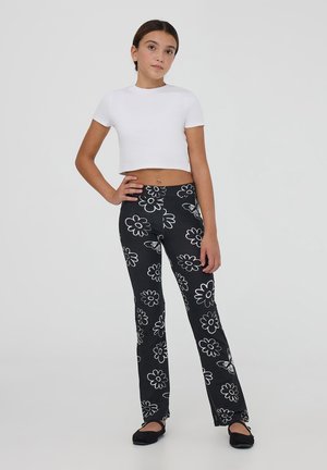 Pantaloni neri a palazzo con motivi floreali bianchi, abbinati a una maglietta bianca cropped. L'outfit presenta una texture liscia e un design semplice.