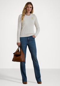 Krem pulover z umetnim kabelskim pletenjem z rumenim logotipom, v kombinaciji z modrimi kavbojkami. Rjava torbica z detajli. S dolgimi rokavi, rebrastimi manšetami in robom.