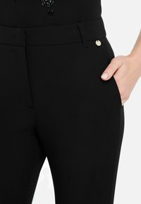 Schwarze maßgeschneiderte Hosen mit glatter Textur, versehen mit einem kleinen goldenen Knopfakzent am Bund und einer Seitentasche, in der eine Hand ruht.