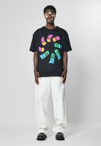 Černé oversized tričko s multicolorovým, hravým textovým designem. Kombinováno s volnými bílými kalhotami a černými botami. Jednoduchý, moderní vzhled.