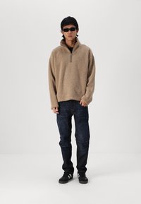 Beige, fuzzy pullover met een quarter-zip kraag, gecombineerd met donkere spijkerbroek. Zwarte zonnebril en sneakers maken de outfit af.