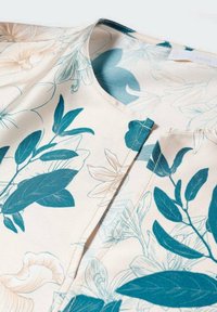 Florale Bluse mit cremefarbenem Hintergrund und türkisen botanischen Mustern. Glatter Stoff, runder Ausschnitt und dezente Nahtdetails.