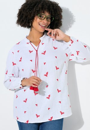 Kvinde iført grønne briller, hvid bluse med røde hjerte mønstre og rød kvast halskæde, smilende og poserer mod hvid baggrund.