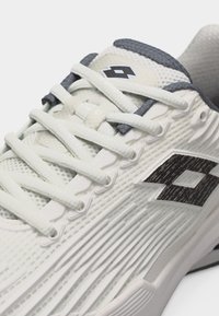 Λευκό αθλητικό παπούτσι με υφή στο πλάι και μαύρο λογότυπο, λευκά κορδόνια και σκουρόχρωμο γκρι επενδεδυμένο περιλαίμιο, εμφανίζεται σε κοντινή λεπτομέρεια.