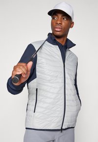 Gai pelēks quilted veste ar tumši zilu piedurknēm, ar augstu apkakli, ar rāvējslēdzēju un sānu kabatām, valkāta ar baltu cepuri un turētām golfa nūjām.