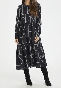 Musta pitkähihaisuus mekko, jossa on valkoisia geometrisia kuvioita, napitettu etuosa ja kerrostettu helma. Valmistettu pehmeästä, teksturoidusta kankaasta.