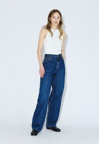 Ung kvinde iført en hvid ærmeløs top, højtaljede vide blå jeans og sorte sko, der står mod hvid baggrund.