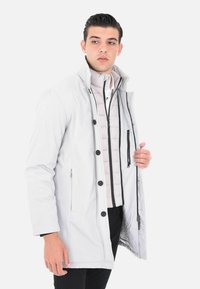 Giacca isolante grigio chiaro con tasche interne con zip, chiusura con bottoni e cappuccio. Presenta uno strato interno gonfiato con una texture a contrasto.