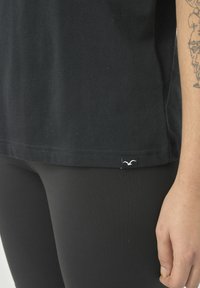 Schwarzes T-Shirt mit kurzen Ärmeln und schlichtem Schnitt, ausgestattet mit einem kleinen weißen Logostick am Saum; getragen mit schwarzen Leggings. Glatte Textur.