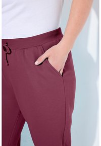 Burgundy tepláky s hladkou texturou, elastickým pasem, šňůrkou a postranními kapsami, předvedené s bílou košili na světlém pozadí.