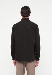 Homme aux cheveux foncés portant une chemise noire à manches longues et un pantalon beige, vu de dos sur un fond clair uni.