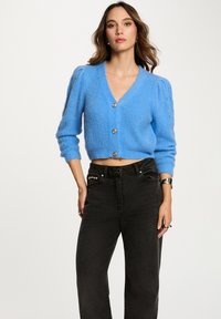 Maglione cropped azzurro chiaro con scollatura a V, texture a coste e bottoni argentati. Presenta maniche puff con motivi ritagliati discreti. Abbinato a jeans neri.