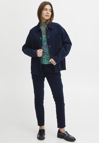 Giacca in corduroy blu navy sopra un top verde a fantasia floreale, abbinata a pantaloni navy coordinati e mocassini neri, che mettono in mostra una vestibilità su misura.