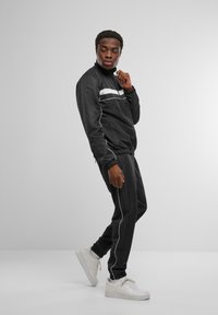 Črni športni trenirki z belimi črtami, narejen iz gladkega materiala. Slim-fit kroj, elastični manšeti in v kombinaciji z belimi supergami.