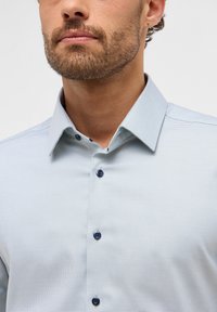 Camicia a scacchi blu chiaro realizzata in tessuto intrecciato, con colletto a bottoni e bottoni blu scuro. Visibili texture e disegno notevoli.