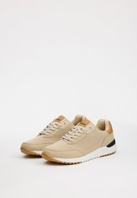 Zapatillas beige con un upper texturizado, acentos en tono marrón claro, suela blanca y una suela de goma. Diseño con cordones y orificios de ventilación.