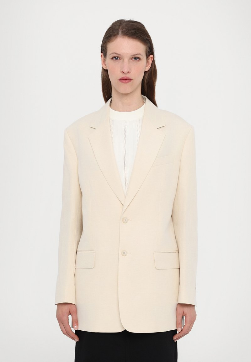 Blazer beige su misura con due bottoni, rever a piega e due tasche frontali. Tessuto liscio e forma strutturata, abbinato a un top bianco.