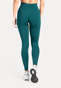 Diepgroene legging met hoge taille, gemaakt van gestructureerde stof en met een aansluitend ontwerp. Draagt witte sportschoenen met een patroon op de zool.