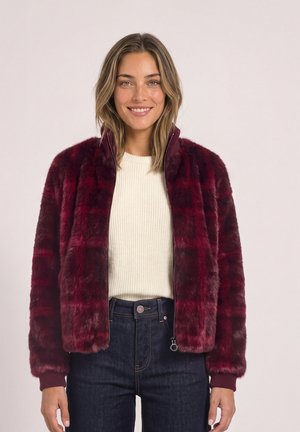Femme portant une veste en fausse fourrure bordeaux sur un pull blanc côtelé et un jean bleu foncé, souriante avec les cheveux lâchés.