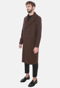 Cappotto doppiopetto marrone con ampio colletto, tasche laterali e tessuto testurizzato. Indossato sopra pantaloni neri aderenti e scarpe nere.