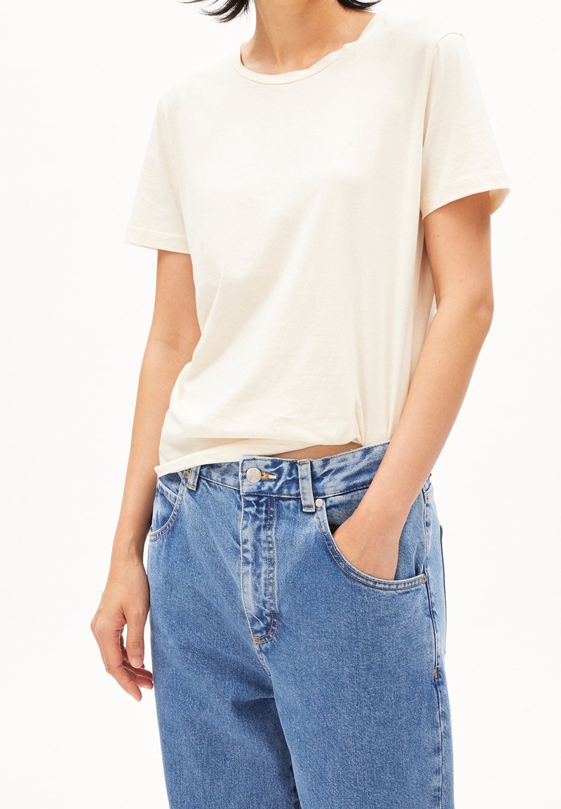 Femme portant un t-shirt blanc uni à manches courtes rentré dans un jean en denim bleu taille haute, avec une main dans la poche.