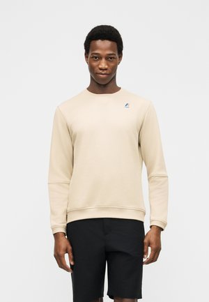 Ung mann iført en beige langermet genser med en liten logo på brystet og svarte shorts, stående mot en ensfarget bakgrunn.