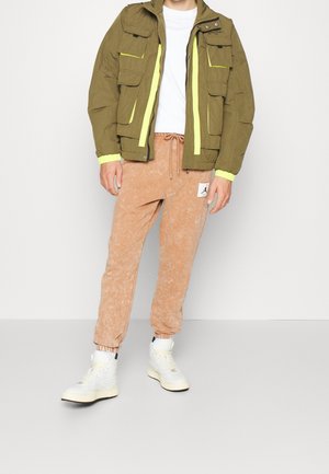 Mann trägt olivgrüne Jacke mit gelben Akzenten, weißes Hemd, sandfarbene Jogginghose mit Kordelzug und weiße High-Top-Sneaker.