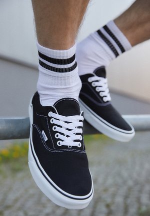 Dwie nogi w białych skarpetkach z czarnymi paskami i czarnych trampkach Vans z białymi sznurówkami odpoczywają na metalowym pręcie na zewnątrz.
