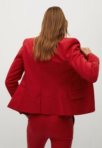 Nainen, jolla on pitkät vaaleanruskeat hiukset, pukeutuneena punaiseen pukuun ja siihen sopiviin punaisiin housuihin, säätää takkia selkäpuolelta luonnollista taustaa vasten.