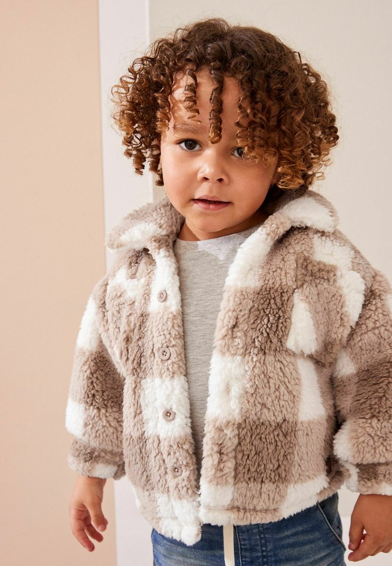 Jeune enfant aux cheveux bouclés portant une veste pelucheuse à carreaux beige et blanc par-dessus un t-shirt gris et un jean bleu.