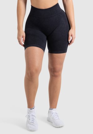 High-waisted zwarte shorts met een gladde textuur en een aansluitend ontwerp, voorzien van een geribde tailleband en zijdelingse paneeldetails. Gecombineerd met witte sneakers.