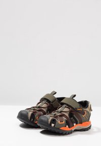 Barnsandaler för utomhusbruk med kamouflage-mönster, tillverkade av mesh och syntetiska material, med justerbara remmar och orange detaljer.