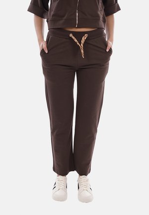 Persona che indossa pantaloni marroni con coulisse e giacca corta, con le mani nelle tasche, abbinati a sneakers bianche con strisce nere.