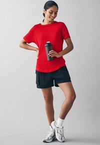 Punane sportlik T-särk lühikeste varrukatega, millele on kombineeritud mustad lühikesed püksid, millel on küljetaskud. Hoidmas tumedat shaker-pudelit, valgetes sokkides ja spordikingades.