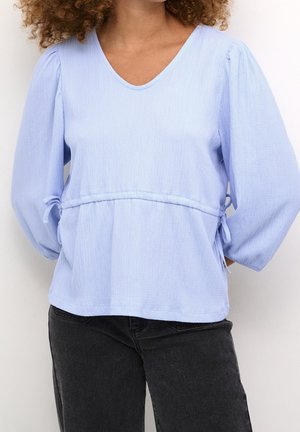 Frau trägt eine hellblaue, strukturierte Bluse mit aufgepufften langen Ärmeln und Seitenschleifen, kombiniert mit schwarzer Jeans.