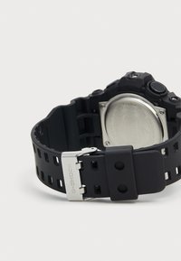 Montre avec bracelet en silicone noir, boîtier hexagonal noir, dos en acier inoxydable et boucle argentée. Comprend plusieurs boutons sur le côté.