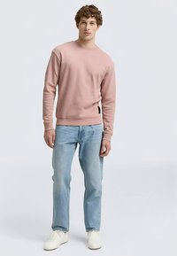 Világos rózsaszín pulóver kerek nyakkivágással, bordázott mandzsettákkal és szegéllyel, világoskék farmerrel és fehér tornacipővel, semleges háttér előtt állva.