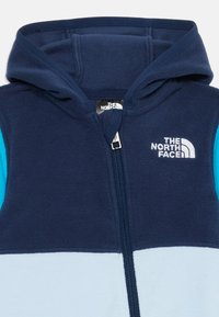 Mornarsko modra in svetlomodra flis jopica s sprednjim zadrgo in vezenim logotipom The North Face na prsih.
