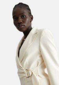 Blazer avorio con dettaglio in vita arricciato, hardware dorato, revers strutturati e texture di tessuto morbido. Vestibilità sartoriale e tasche laterali.