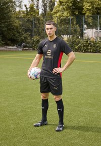 Black football jersey with red accents, black shorts, and black socks. Player holds a multicolored soccer ball on green turf field.