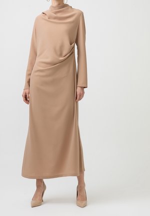 Femme portant une robe midi beige à manches longues et col haut avec un détail drapé sur le devant, associée à des escarpins beiges à bout pointu.