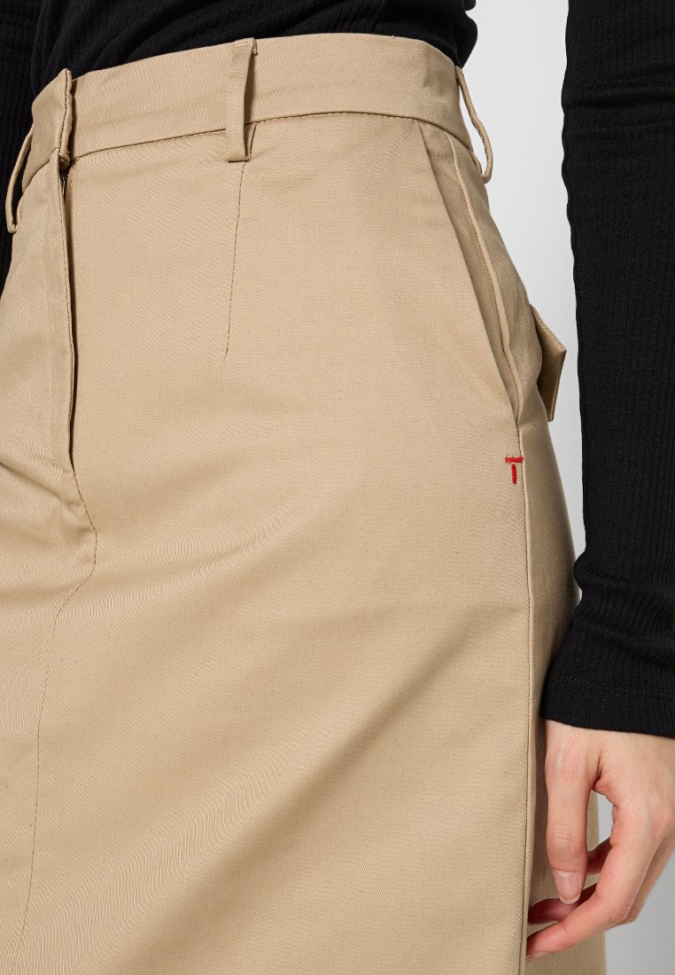 Beżowa spódnica z teksturowanej tkaniny, z bocznymi kieszeniami, z paskiem w talii i małym czerwonym logo z przodu.